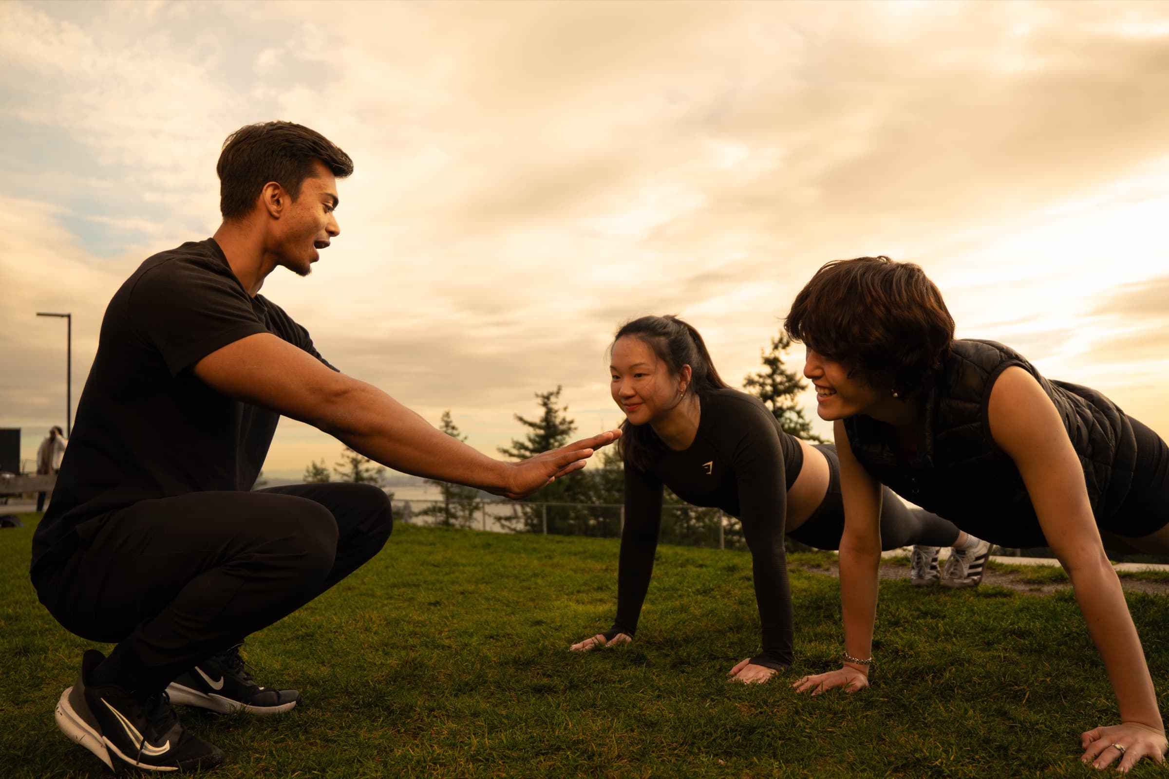 A coach crouched beside two clients holding a plank on grass at golden hour